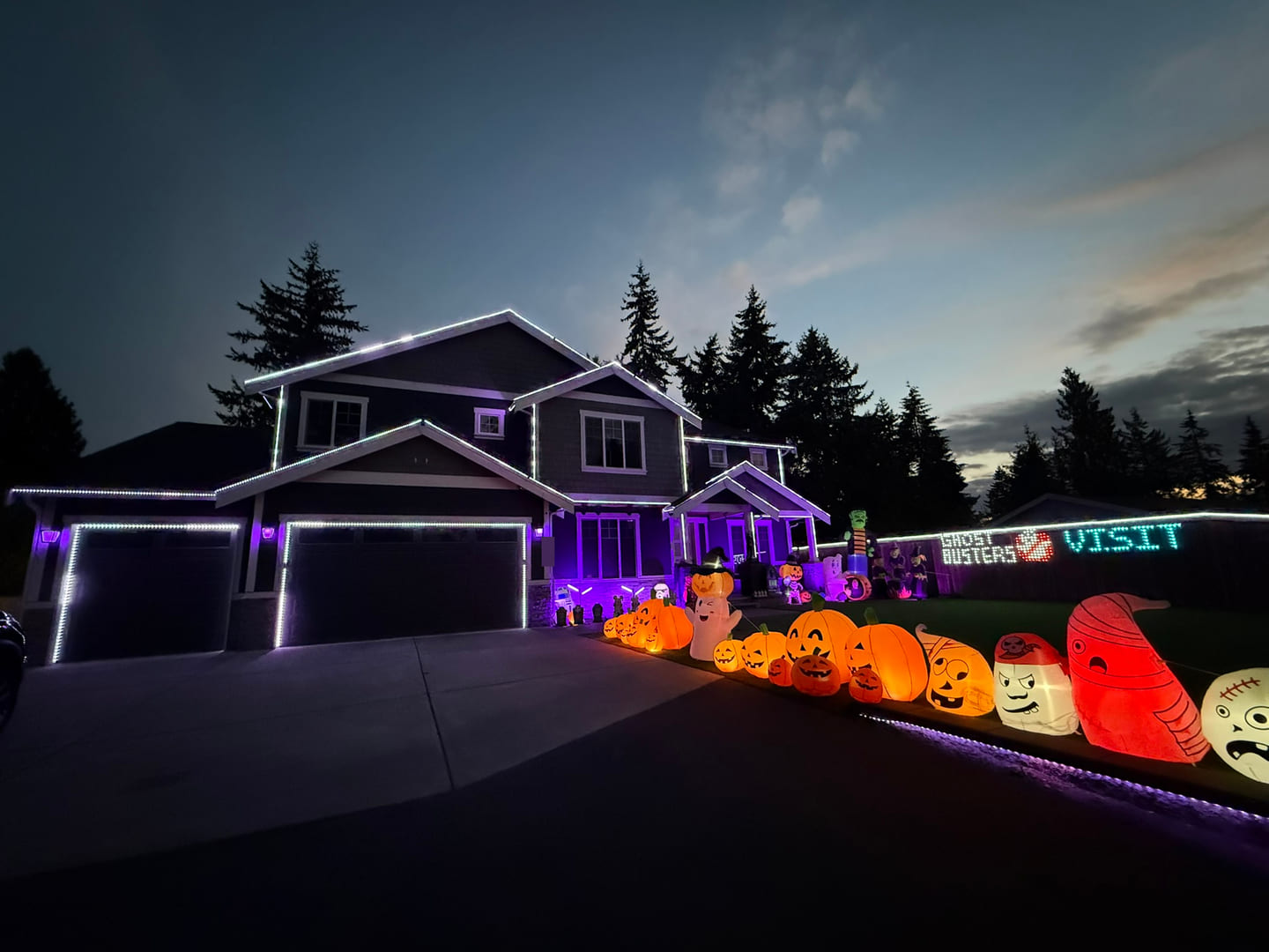 A row of orange Halloween pumpkins and character inflatables beside the driveway, with the illuminated House With The Lights roofline to the left