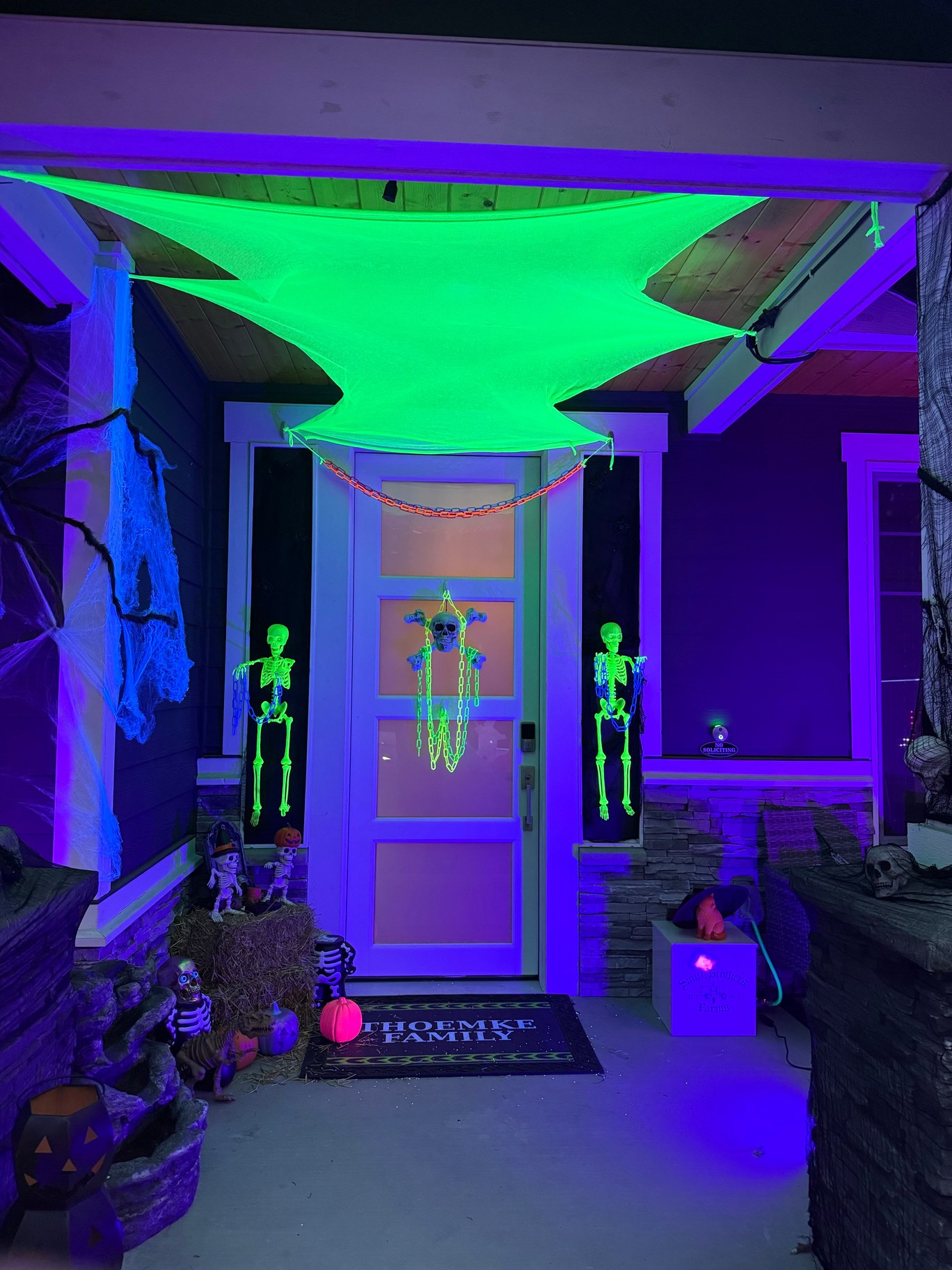 Halloween porch glowing purple and green under blacklights, with fluorescent fabric overhead, hanging skeletons, cobwebs, pumpkins, and a decorated front door.