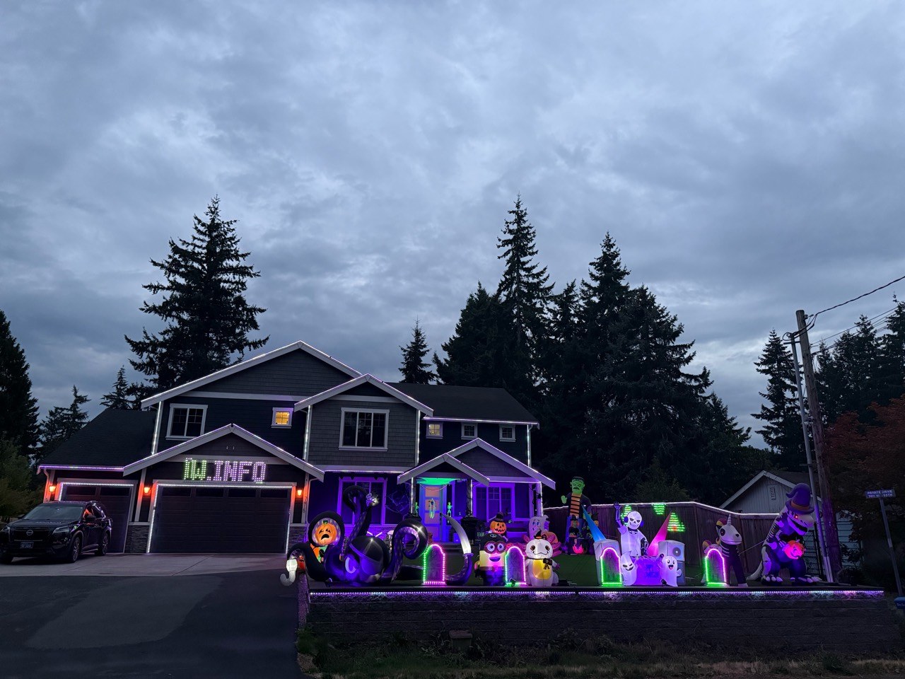 House With The Lights at dusk, with four illuminated tombstones along the retaining wall, Halloween inflatables, a net light above the garage, and purple porch lighting.