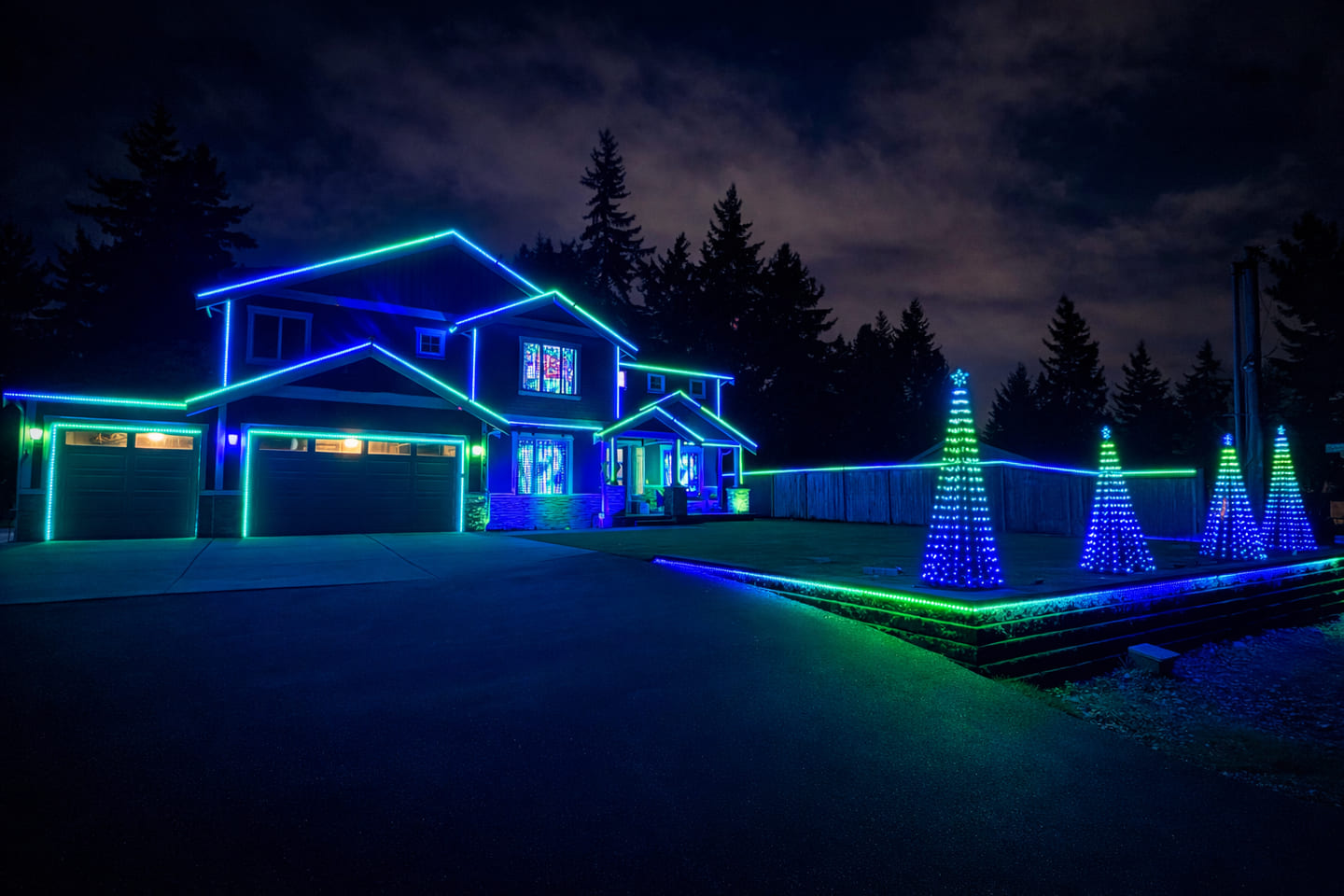 House With The Lights illuminated in blue, cyan, and green beneath a cloudy night sky, with lighted cone trees along the front lawn