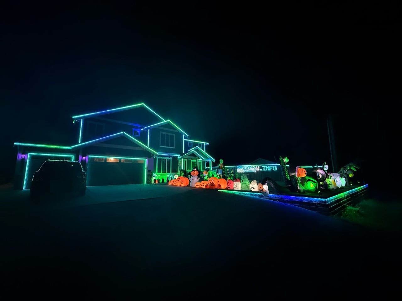 House With The Lights at night, with blue and green lights outlining the roof, garage doors, and yard above glowing Halloween inflatables