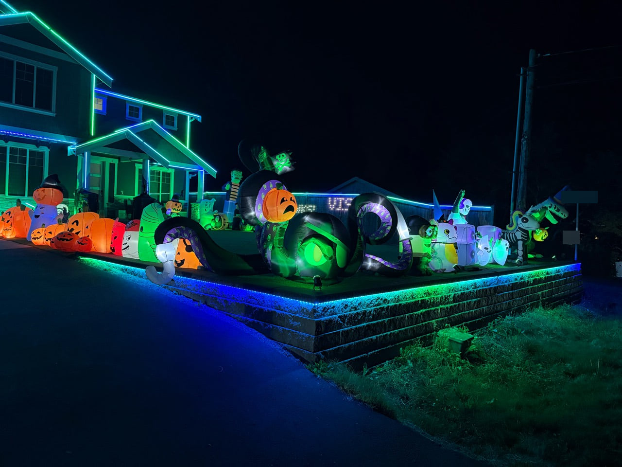 Blue and green lights trace the front wall and porch around pumpkins, ghosts, skeletons, and a large tentacled Halloween inflatable holding a pumpkin